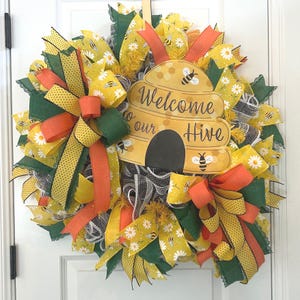 May include: A yellow and green wreath with a beehive sign that says "Welcome to our Hive". The wreath is decorated with yellow and orange ribbons, white daisies, and black and yellow bees.