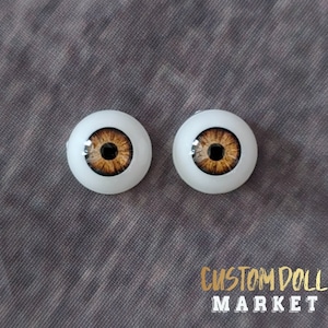 May include: Two doll eyes with a light brown iris and a dark brown pupil, set in a white, rounded base. The eyes are displayed on a gray fabric surface. The text "CUSTOM DOLL MARKET" is visible in gold.