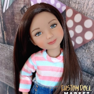 Op de afbeelding: Een op maat gemaakte pop met lang bruin haar, blauwe ogen en een roze en blauw gestreept shirt. De pop draagt een spijkerbroek. Het Custom Doll Market-logo is zichtbaar.