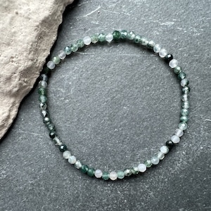 Könnte beinhalten: Ein filigranes Armband aus kleinen, facettierten Perlen in Grün, Weiß und Schwarz. Die Perlen sind zu einem Kreis aufgefädelt. Das Armband wird auf einer dunkelgrauen Oberfläche neben einem hellgrauen Stein gezeigt.