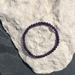 Könnte beinhalten: Ein zartes Amethyst-Perlenarmband. Das Armband besteht aus kleinen, runden, tiefvioletten Perlen. Das Armband liegt auf einer strukturierten, cremefarbenen Oberfläche, mit einem dunkelgrauen Hintergrund.