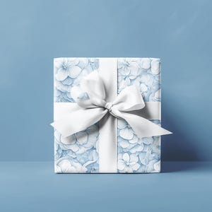 Puede incluir: Una caja de regalo cuadrada envuelta en papel estampado floral azul claro. Una cinta ancha de sat&eacute;n blanco est&aacute; atada alrededor de la caja con un lazo grande en la parte superior. El fondo es azul claro liso.
