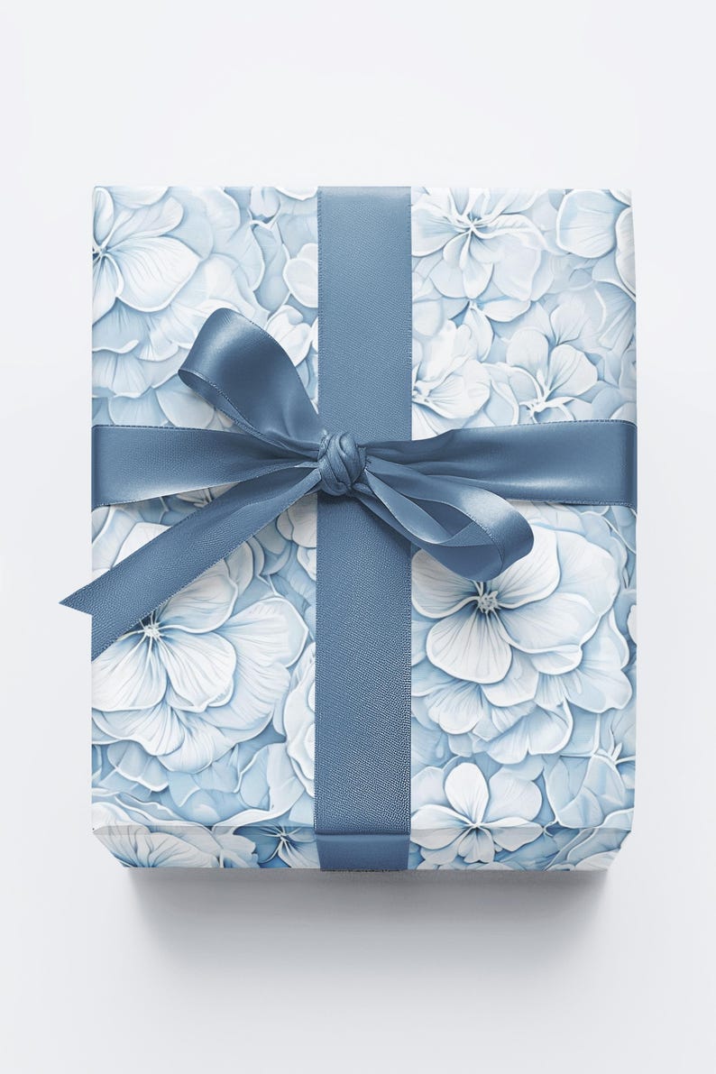 Puede incluir: Un regalo envuelto en papel con un estampado de flores de hortensia blancas y azul claro. Una cinta y un lazo azules a juego adornan el regalo. La imagen sugiere un tema de celebraci&oacute;n y regalos, adecuado para diversas ocasiones.