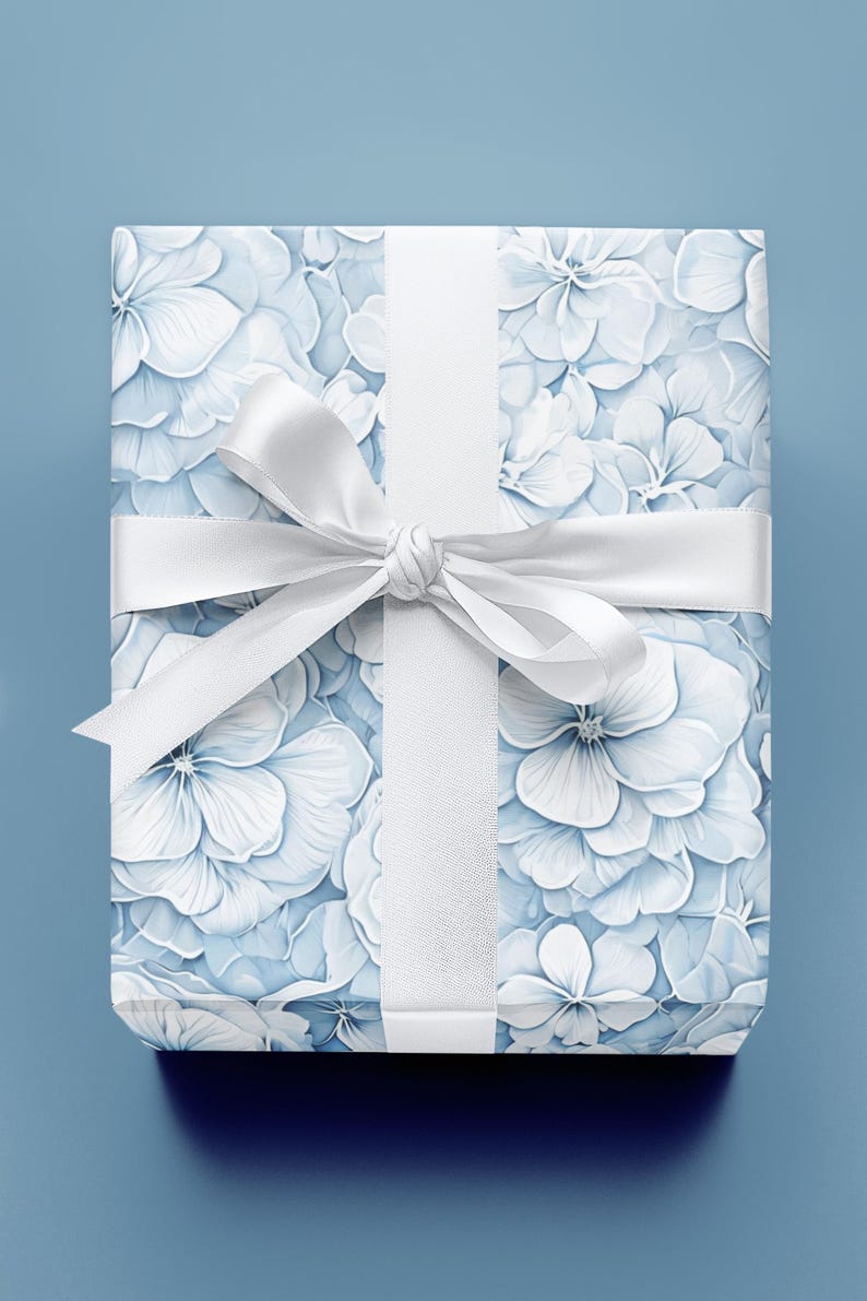 Puede incluir: Un regalo envuelto con un estampado floral de flores de hortensia blancas y azul claro. El regalo est&aacute; atado con una cinta y un lazo blancos, sobre un fondo azul claro.