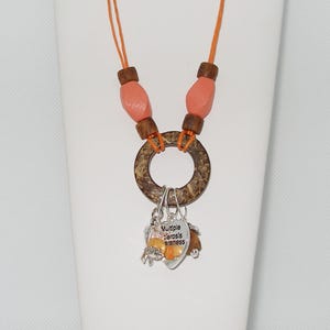 May include: A necklace with an orange cord, wooden beads, and a large circular pendant. The pendant has a silver charm with the words "Multiple Sclerosis Awareness" and other charms with orange beads.