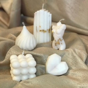 May include: Five white candles of different shapes and sizes. The candles are arranged on a beige fabric surface. The candles are made of wax and have a smooth, matte finish.