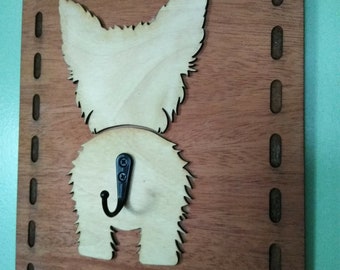 May include: Wooden wall-mounted hook featuring a light-colored dog silhouette with a black hook. The dog design is set against a rectangular, dark brown wooden background with a decorative border. The hook is ideal for hanging items.