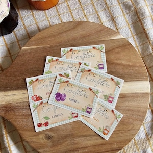 Puede incluir: Una tabla de cortar de madera muestra varias etiquetas para mermeladas caseras. Las etiquetas, con el texto "FAIT MAISON Confiture", presentan ilustraciones de frutas como cerezas, ciruelas y albaricoques. Se ven tarros de mermelada en el fondo.