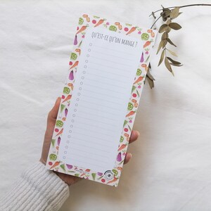 Könnte beinhalten: Ein weißer Notizblock mit einem linierten Innenraum und einem Rand aus bunten Gemüse. Der Notizblock hat den Text "Qu'est-ce qu'on mange?" oben aufgedruckt.