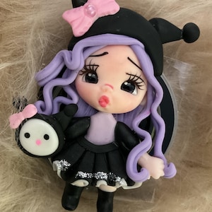 May include: A small figurine featuring a character with long wavy lavender hair, wearing a black jester-style hat with a pink bow and a black dress with silver glitter accents. The character holds a small round black and white accessory.
