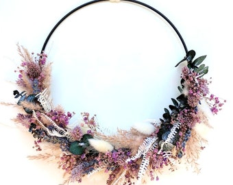 May include: A dried floral wreath with a black metal hoop. The wreath is decorated with dried flowers in shades of purple, pink, and white, as well as dried grasses and eucalyptus.