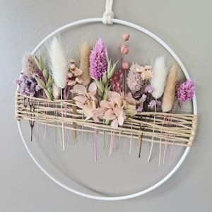 Könnte beinhalten: Ein kreisförmiger floraler Wandbehang mit getrockneten Blumen in Rosa, Lila und Beige. Die Blumen sind in einem weißen Metallreifen angeordnet, mit einer gewebten Basis und einer Aufhängeschnur. Die Anordnung umfasst flauschiges Pampasgras und zarte Blüten.