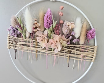May include: A circular floral wall hanging with dried flowers in pinks, purples, and beige. The flowers are arranged within a white metal hoop, with a woven base and hanging cord. The arrangement includes fluffy pampas grass and delicate blooms.
