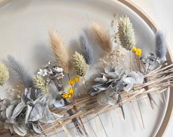 Puede incluir: Un arreglo de flores secas en un marco de madera circular. El arreglo presenta hierbas secas, flores y hojas en tonos grises, beige y amarillos.