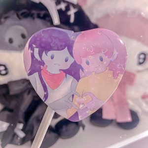 May include: A heart-shaped pin featuring two cartoon characters holding hands. The character on the left has purple hair and is wearing a red bandana. The character on the right has pink hair and is wearing a yellow shirt. The pin has a glittery finish.