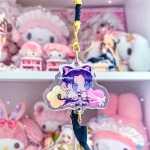 May include: A clear acrylic keychain featuring a chibi anime character with purple hair, wearing a black and white maid outfit with a cat ear headband. The character is holding a pink and white heart-shaped object. The keychain has a black cord and a gold metal clasp.
