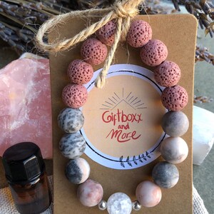 May include: A pink and gray beaded bracelet with a white crystal charm. The bracelet is on a brown card with the text "Giftboxx and More".