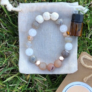 May include: A beaded bracelet with a variety of stone beads in shades of white, gray, and peach, accented with gold-colored metal beads. The bracelet rests on a small, beige drawstring bag, next to a small brown essential oil bottle.