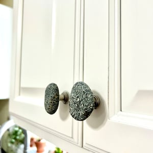 May include: Close-up of two gray stone cabinet knobs with silver hardware. The knobs are oval shaped and have a rough, textured surface.