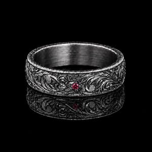 Puede incluir: Un anillo de metal gris oscuro con un intrincado patrón floral en relieve en toda la banda exterior. Una pequeña piedra preciosa redonda de color rojo vibrante está engastada en el centro del grabado detallado del anillo.