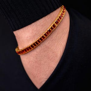 May include: A gold-toned bracelet with rectangular, amber-colored stones. The bracelet is worn on a person's wrist, with a black shirt in the background. The bracelet's design is a continuous line of stones.