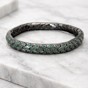 May include: A dark gray bracelet with a honeycomb design. Each cell of the honeycomb is filled with a small, emerald-green gemstone. The bracelet is set against a white and gray marble surface.