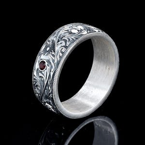 May include: A silver band ring featuring intricate, dark-oxidised floral scrollwork engravings across the outer surface. The ring is accented with a single, small, round, deep red gemstone set within the detailed metalwork pattern.