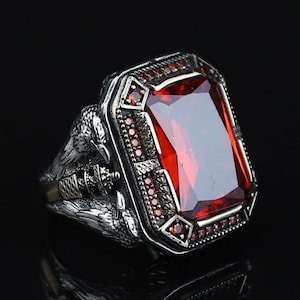 May include: A silver ring featuring a large, rectangular, faceted red stone set in a frame accented with small red stones. The band is detailed with sculpted bird motifs on both sides, showcasing intricate metalwork and a dark, oxidised finish.
