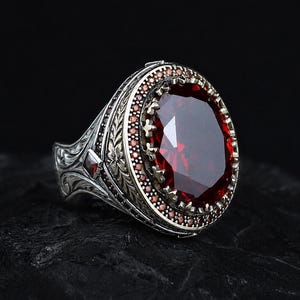 Könnte beinhalten: Ein Silberring mit einem großen, oval geschliffenen, tiefroten Edelstein in der Mitte. Die Fassung ist von einer Umrandung aus winzigen roten Steinen und aufwendigen Metallgravuren entlang der Ringschiene mit detaillierten Blumenmustern umgeben.