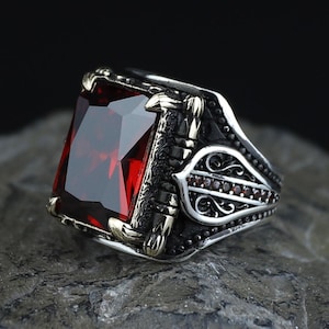 Pode incluir: Um anel de prata com uma grande pedra preciosa retangular facetada de cor vermelho-escura, inserida numa armação de metal em forma de garra. A banda apresenta padrões intrincados de filigrana de prata oxidada escura e gravações decorativas.