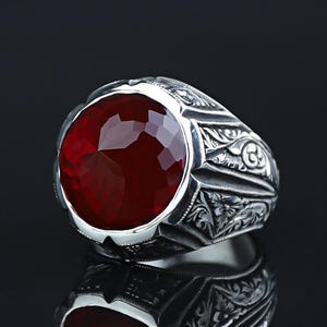 Könnte beinhalten: Ein silberner Ring mit einem großen, facettierten, tiefroten Edelstein. Das Ringband weist aufwendige, eingravierte Blumenmuster auf. Der Edelstein ist oval und fängt das Licht ein. Der Ring wird auf einer reflektierenden Oberfläche präsentiert.