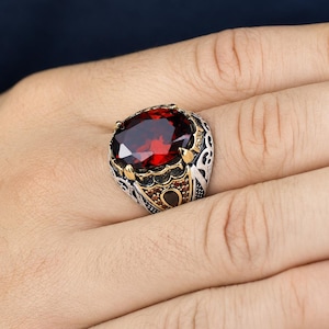 May include: A silver ring featuring a large, oval-cut, deep red gemstone set in a pronged mounting. The band is decorated with intricate, dark-oxidized metalwork, gold-toned accents, and small, embedded amber-colored stones along the sides.
