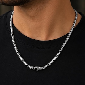 Puede incluir: Un collar de cadena en tono plateado con un patrón de eslabones texturizados de estilo tejido. El collar incluye un elemento de cierre cilíndrico en la parte delantera, acentuado con pequeñas piedras circulares oscuras en cuadrícula.