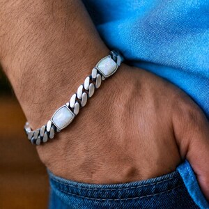 May include: A silver-toned curb chain bracelet featuring two rectangular, iridescent white opal-like stones set within the metal links. The bracelet has a polished finish and a classic interlocking link design that wraps around the wrist.