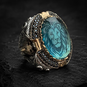 May include: A silver and gold ring with a large, oval, blue gemstone featuring a lion's head engraving. The ring has intricate detailing, including black accent stones and a lion's head on the side.