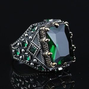 Könnte beinhalten: Ein silberfarbener Ring mit einem großen, rechteckigen, facettierten dunkelgrünen Stein in der Mitte. Die Ringschiene ist mit kleineren grünen Steinen und aufwendigen Metallarbeiten verziert, darunter erhabene Noppen und eine dunkle Textur.