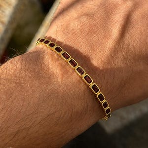 May include: A gold-colored bracelet with rectangular, dark red gemstones. The bracelet is worn on a wrist, showcasing the jewelry's design and color contrast. The gemstones are set in individual gold frames.