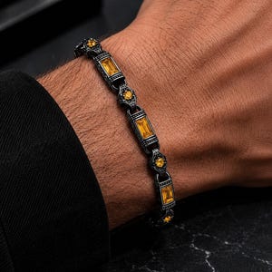 May include: A dark gunmetal-toned bracelet featuring alternating rectangular and circular links. The rectangular links are set with baguette-cut yellow stones, while the circular links contain smaller round-cut yellow stones in a detailed setting.