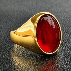 May include: A gold-toned ring featuring a smooth, polished band and a large, oval-shaped, deep red cabochon stone set securely in the center. The metal surface has a reflective finish, and the red stone displays a translucent, glossy appearance.