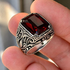 Peut inclure: Une bague en argent ornée d'une grande pierre précieuse rectangulaire à facettes rouge foncé, sertie dans un cadre à bordure perlée. L'anneau présente des motifs complexes en relief de volutes et de feuilles sur les côtés du sertissage.