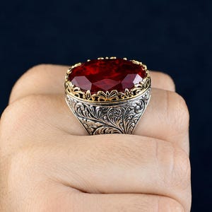 May include: A ring featuring a large, oval-cut, deep red gemstone set in a gold-toned filigree bezel. The band is crafted from oxidised silver with intricate, raised floral scrollwork engravings that wrap around the entire outer surface.