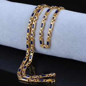 Pode incluir: Colar dourado com detalhes azuis retangulares e redondos. A corrente está sobre uma superfície cinza clara, mostrando o design intrincado e o contraste de cores. O colar possui um fecho.