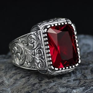 Pode incluir: Um anel de prata com uma grande pedra preciosa retangular de cor vermelho-escura facetada, engastada numa moldura com bordas entalhadas. A banda apresenta intrincados gravados florais em espiral que se estendem pelos lados do engaste.