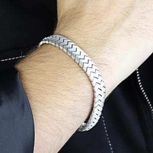 May include: A silver bracelet with a chevron pattern is worn on a wrist. The bracelet is made of interlocking metal segments. The person is wearing a black jacket.