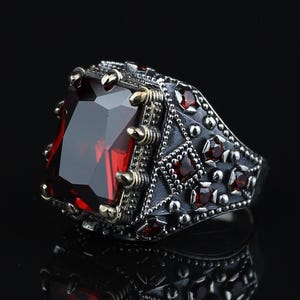 Puede incluir: Un anillo de tono plateado con una gran piedra preciosa roja rectangular facetada sujeta por garras. La banda está decorada con un patrón de celosía de metal oscuro texturizado y piedras rojas cuadradas más pequeñas incrustadas.