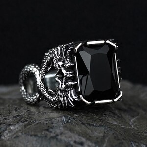 May include: A silver-toned metal ring featuring a large, rectangular, faceted black stone set in the center. The band is intricately detailed with a textured, scaled serpent design that wraps around the sides of the central stone setting.