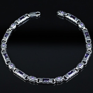 Puede incluir: Una pulsera de plata con piedras preciosas moradas rectangulares y en forma de diamante. La pulsera está hecha de eslabones interconectados con detalles ornamentados. El cierre está abierto. Fondo negro.