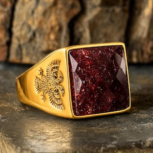 Op de afbeelding: Een goudkleurige ring met een grote, vierkante, facetgeslepen, donkerrode steen. De ring heeft aan één kant een reliëf adelaarsontwerp. De ring ligt op een donker, getextureerd oppervlak.