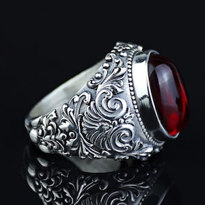 May include: A silver ring with an oval, deep red gemstone. The ring has detailed scrollwork and floral patterns. The gemstone is set in a polished silver bezel. The ring is displayed on a reflective black surface.