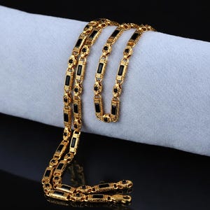 May include: A gold-colored chain necklace with black rectangular and round accents. The necklace is draped over a light gray surface, with a black background. The chain has a repeating pattern of rectangular black stones.